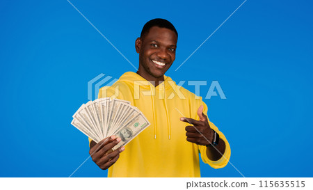 Cheerful young black guy points finger at many money dollars, isolated on blue background, studio, panorama. Recommendation finance, human emotions, profit, victory and wealth Cheerful young black guy points finger at many money dollars, isolated on blue background, studio, panorama. Recommendation finance, human emotions, profit, victory and wealth 115635515