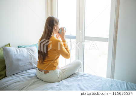 A teen girl sits on a neatly made bed, wearing a yellow sweater and white pants, drinking a cup of coffee as she gazes out a large window, enjoying the bright morning light streaming into the room. 115635524