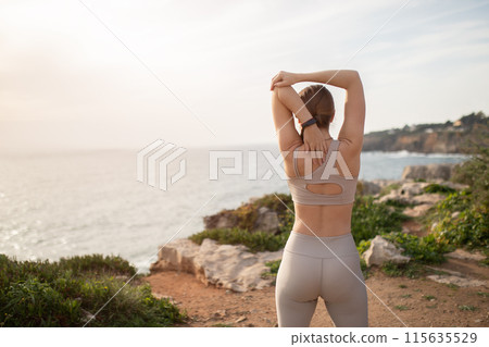 Millennial caucasian lady athlete in sportswear and fit tracker doing arm stretching, enjoy workout, exercises at sea beach, back. Fitness outdoor, sports, body care alone and active lifestyle 115635529