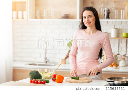 A woman is standing in a kitchen, focused on chopping a variety of colorful vegetables on a wooden cutting board. She holds a sharp knife in one hand and skillfully slices veggies 115635531