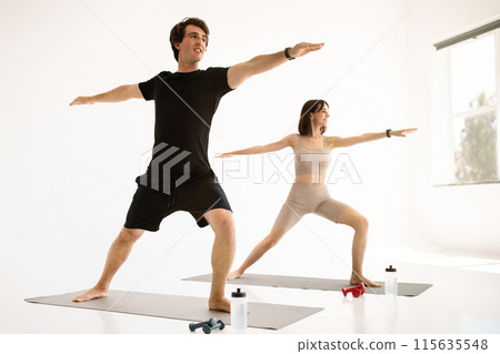 Happy young european guy and woman in sportswear doing hand exercises on mat in living room or gym interior. Water for health care, dumbbells for body care, workout together, sports lifestyle Happy young european guy and woman in sportswear doing hand exercises on mat in living room or gym interior. Water for health care, dumbbells for body care, workout together, sports lifestyle 115635548