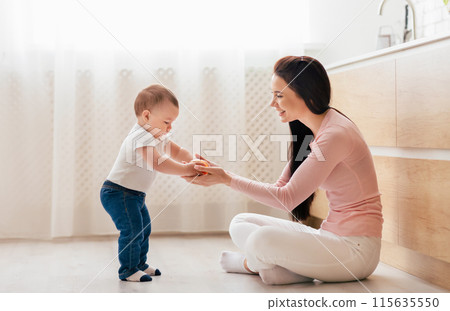 A mother sits on the kitchen floor, encouraging her baby, who is taking tentative steps towards her. The morning light filters through the sheer curtains, creating a warm and cheerful atmosphere. A mother sits on the kitchen floor, encouraging her baby, who is taking tentative steps towards her. The morning light filters through the sheer curtains, creating a warm and cheerful atmosphere. 115635550