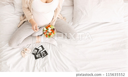 A woman is seated on a bed baby ultrasound image and booties, holding a plate of food in her hands. She appears to be enjoying her meal in a relaxed setting, top view, copy space A woman is seated on a bed baby ultrasound image and booties, holding a plate of food in her hands. She appears to be enjoying her meal in a relaxed setting, top view, copy space 115635552