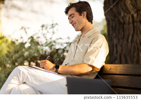 Millennial man student sitting on bench with computer laptop by him, taking notes in notepad, doing homework while enjoying warm sunny day at park. Happy guy studying online via pc outdoor 115635553