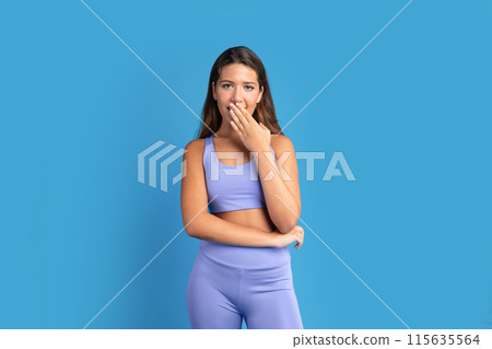 Glad shocked slim young caucasian lady in sportswear covers open mouth with hand after workout, isolated on blue studio background. Surprise emotions, good news, fitness, body care and weight loss 115635564