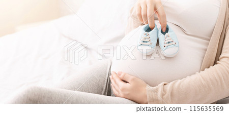 Millennial woman playing with her belly, trying tiny baby shoes on it, close up, panorama with free space 115635569