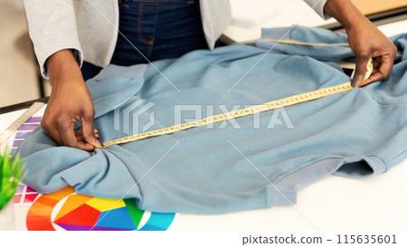 African American woman measuring a shirt using a tape measure. The individual is carefully extending the tape measure along the length and width of the cloth to determine its dimensions accurately. 115635601