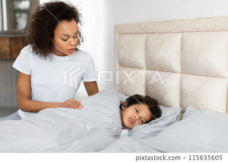 A concerned African American young woman is standing beside a bed, gently looking down at a child who appears to be unwell and resting under a cozy blanket 115635605