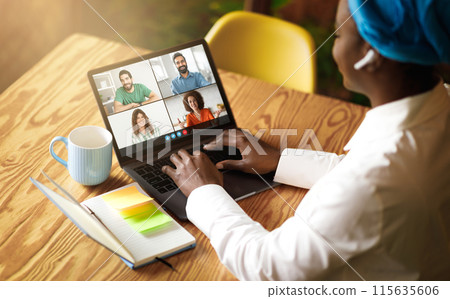 African American woman is sitting at a wooden table, engaging in a virtual meeting on her laptop with four colleagues. She has a colorful book and a mug beside her. 115635606