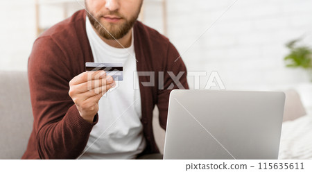 A man is pictured holding a credit card in one hand and looking at a laptop screen with a focused expression. He appears to be making an online purchase or managing his finances, cropped A man is pictured holding a credit card in one hand and looking at a laptop screen with a focused expression. He appears to be making an online purchase or managing his finances, cropped 115635611