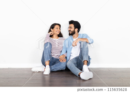Excited thrilled beautiful young indian couple sitting on floor at new empty apartment over white blank wall background, holding hands, have conversation, dreaming about future house, copy space Excited thrilled beautiful young indian couple sitting on floor at new empty apartment over white blank wall background, holding hands, have conversation, dreaming about future house, copy space 115635628