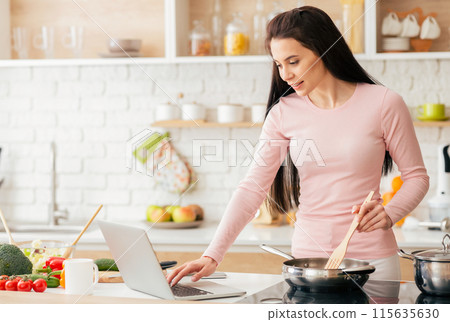A woman is multitasking in the kitchen, cooking food while using a laptop for work or browsing the internet. She is standing near the stove, focusing on both tasks at hand. A woman is multitasking in the kitchen, cooking food while using a laptop for work or browsing the internet. She is standing near the stove, focusing on both tasks at hand. 115635630