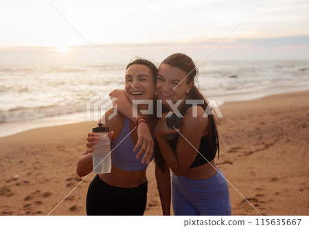 Laughing smiling millennial caucasian twins sisters women hugging, have fun with bottle of water, enjoy sports together on sea beach. Morning workout, health care, active and motivation Laughing smiling millennial caucasian twins sisters women hugging, have fun with bottle of water, enjoy sports together on sea beach. Morning workout, health care, active and motivation 115635667
