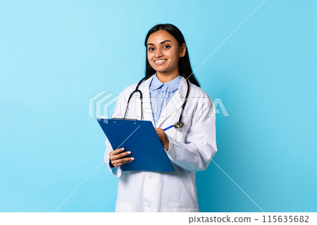 Medical Offer. Confident Young Indian Doctor Lady In White Coat Uniform Posing With Stethoscope And Clipboard, Smiling At Camera, Standing Over Blue Background. Healthcare, General Practitioner 115635682