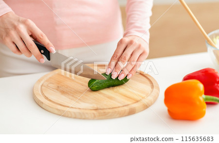 Lady uses a knife to slice a cucumber on a wooden cutting board in a brightly lit kitchen. Nearby on the counter, red and yellow bell peppers are also visible. 115635683