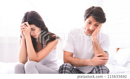 A couple sits on a white bed with sad expressions on their faces. The woman is wearing a gray tank top and pants, and the man is wearing a white t-shirt and plaid pants. 115635692