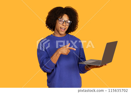 A young man wearing a blue sweater and glasses points at his laptop. The background is a vibrant orange, creating a playful and energetic contrast. 115635702