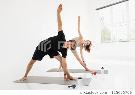 Cheerful millennial caucasian couple in sportswear training together, practicing yoga with dumbbells and bottle of water in living room, gym interior. Workout, active lifestyle, sport and fitness Cheerful millennial caucasian couple in sportswear training together, practicing yoga with dumbbells and bottle of water in living room, gym interior. Workout, active lifestyle, sport and fitness 115635706