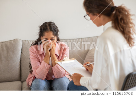 A woman is sitting on a couch in an office, crying while being counseled by a therapist. The therapist is sitting to her right, holding a clipboard with a pen in her hand, seemingly taking notes. 115635708