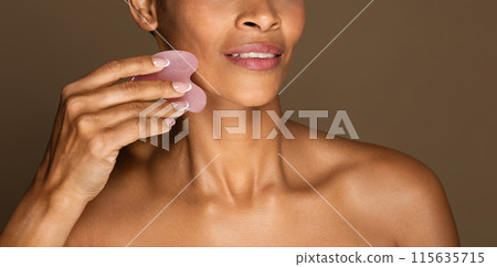 Aged skin lifting. Black middle aged woman massaging face with gua sha tool, making anti-aging skin care routine with jade guasha stone scraper, panorama, cropped, closeup Aged skin lifting. Black middle aged woman massaging face with gua sha tool, making anti-aging skin care routine with jade guasha stone scraper, panorama, cropped, closeup 115635715