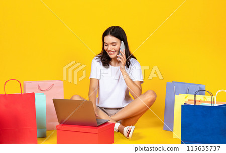 Happy young woman with shopping bags and laptop talking on cellphone, smiling shopaholic lady making online purchases, confirming web order, buying in internet, sitting on yellow studio background 115635737