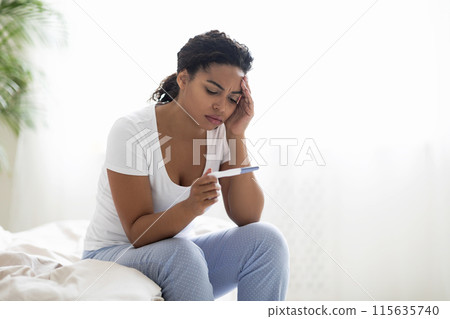 Portrait Of Upset Young Black Female With Pregnancy Test In Hand Sitting On Bed, Upset African American Woman Suffering Health And Fertility Problems, Touching Head And Frowning, Relaxing In Bedroom 115635740