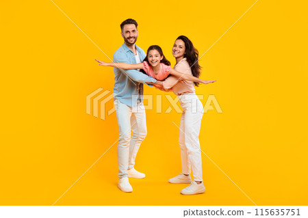 Happy parents holding daughter, girl spreading arms like plane, posing and having fun, smiling to camera over yellow background, full length, free space 115635751