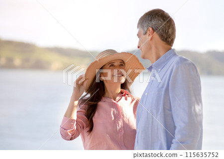 Romantic Mature Spouses Having Walk Near Sea In Summer Time, Loving Middle Aged Couple Relaxing Outdoors And Bonding With Each Other, Wife Looking At Husband With Love, Wearing Straw Hat 115635752