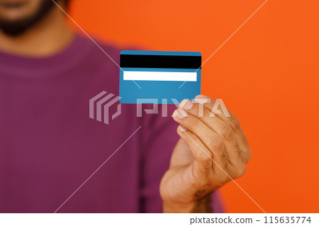 Banking concept. Cropped image of young dark-skinned man wearing basic casual t-shirt holding in hand credit bank card isolated on bright orange colour background, studio shot, blurred, copy space 115635774