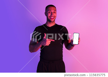 Cheerful African American Young Man Showing Phone With Empty Screen In Purple Neon Lighting In Studio. Guy Wearing Sportswear Pointing At Smartphone Showing Gadget, Mockup For Workout App Advert 115635775