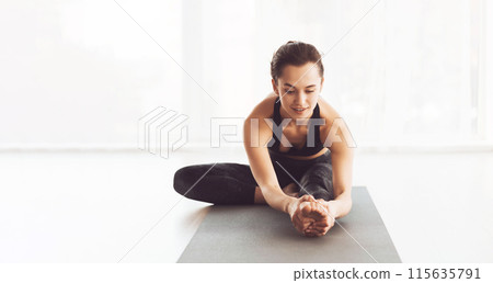 A young woman is practicing a yoga stretch indoors on a mat during the morning. She is wearing comfortable workout attire and appears focused and calm. 115635791