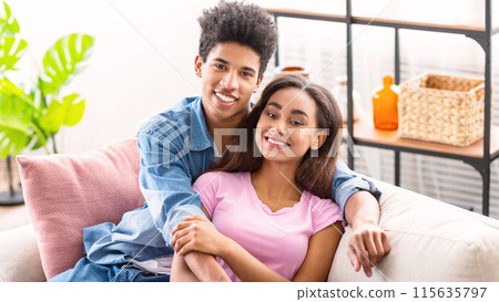 Black teen couple relaxing together on a couch in a living room. The guy has his arm around the girl, and they are both smiling and looking at the camera. 115635797