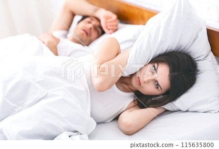 A woman lies in bed, covering her ears with a pillow, while her partner sleeps soundly beside her. She appears frustrated or annoyed, possibly due to her partners snoring. A woman lies in bed, covering her ears with a pillow, while her partner sleeps soundly beside her. She appears frustrated or annoyed, possibly due to her partners snoring. 115635800