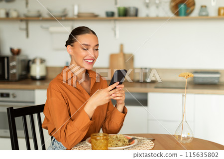 Cheerful lady using smartphone while sitting at kitchen table and having lunch, texting on mobile phone, surfing internet at home, free space 115635802