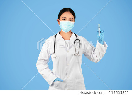 Immunization Concept. Confident asian female doctor wearing protective medical mask holding syringe with vaccine and looking at camera, professional medical worker posing over blue studio background 115635821