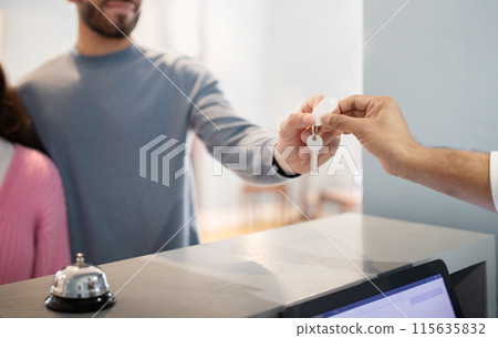 Unrecognizable Travelers Couple Checking In At Hotel Taking Accommodation Room Key From Receptionist Man, Standing At Reception Counter Indoor. Cropped Shot, Selective Focus Unrecognizable Travelers Couple Checking In At Hotel Taking Accommodation Room Key From Receptionist Man, Standing At Reception Counter Indoor. Cropped Shot, Selective Focus 115635832
