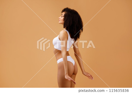 Young slim black lady with perfect ideal body shape posing isolated on beige studio background, half-turned sexy slender woman in white underwear, free space Young slim black lady with perfect ideal body shape posing isolated on beige studio background, half-turned sexy slender woman in white underwear, free space 115635836
