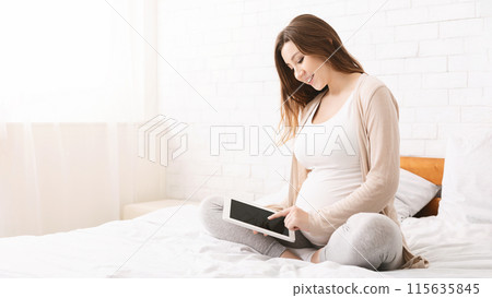 A pregnant woman is sitting cross-legged on a bed and smiling while using a tablet, creating a peaceful atmosphere as she enjoys her time connected to her device, copy space A pregnant woman is sitting cross-legged on a bed and smiling while using a tablet, creating a peaceful atmosphere as she enjoys her time connected to her device, copy space 115635845