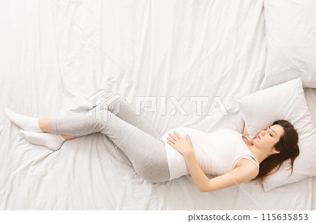 A pregnant woman is reclining on a bed surrounded by soft pillows. She appears comfortable and supported as she rests, top view, copy space 115635853