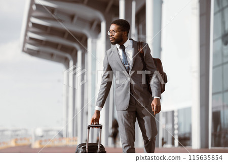 Black businessman walking with rolling suitcase outside a modern airport, ready for a business trip or travel. 115635854
