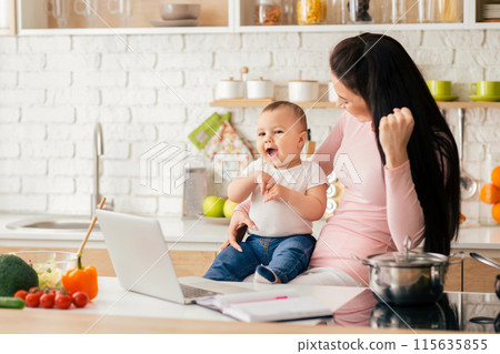 A mother and baby share a joyful moment in a modern kitchen. The baby sits on the counter while the mother looks at a laptop, both smiling and engaged. 115635855