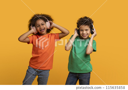 Two African American children stand in front of a yellow background while listening to music with headphones 115635858