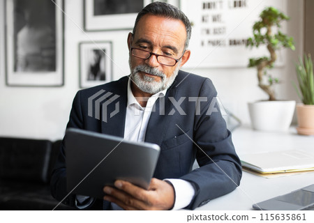 Serious handsome caucasian senior businessman in suit and glasses uses tablet in modern office interior, close up. Finance, work remotely, online business with app and startup Serious handsome caucasian senior businessman in suit and glasses uses tablet in modern office interior, close up. Finance, work remotely, online business with app and startup 115635861