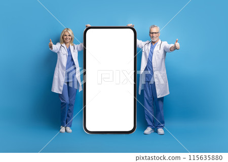 Friendly cheerful senior doctors man and woman in medical white coats showing great mobile app for telemedicine, poisng by phone with white blank screen, showing thumb up, blue background, mockup Friendly cheerful senior doctors man and woman in medical white coats showing great mobile app for telemedicine, poisng by phone with white blank screen, showing thumb up, blue background, mockup 115635880