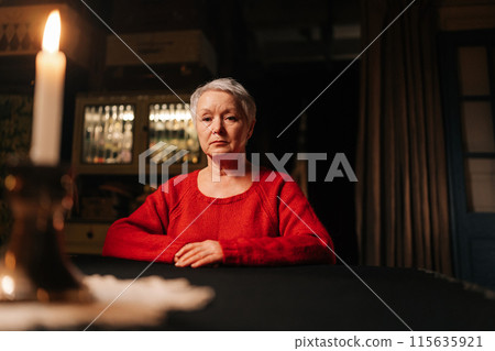 Low-angle view portrait of gray-haired middle-aged woman in casual red sweater looking at camera with serious expression while sitting at table in dark room, by light of burning candle. 115635921