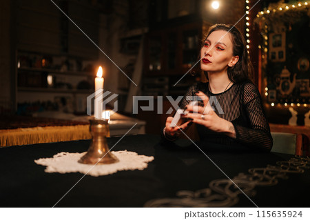 Confident mysterious fortuneteller shuffling deck of tarot cards by light of burning candles, looking away with serious expression. Sorceress shuffles Tarot cards before forecasting future for client Confident mysterious fortuneteller shuffling deck of tarot cards by light of burning candles, looking away with serious expression. Sorceress shuffles Tarot cards before forecasting future for client 115635924