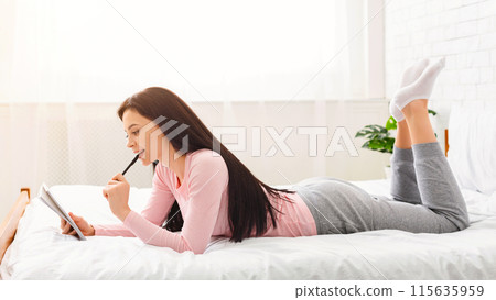 A young woman is captured lying on her stomach on a bed, with her feet playfully raised in the air. She is focused on writing something in a notebook with a pen, wearing casual indoor clothing A young woman is captured lying on her stomach on a bed, with her feet playfully raised in the air. She is focused on writing something in a notebook with a pen, wearing casual indoor clothing 115635959