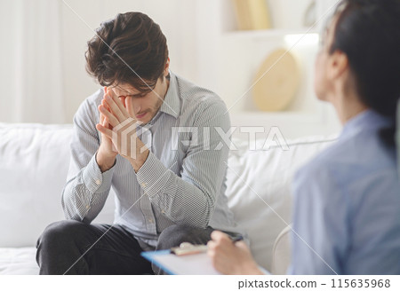 A distressed man sits on a couch with his head in his hands, engaging in a conversation with a counselor who takes notes. The setting appears to be a bright 115635968