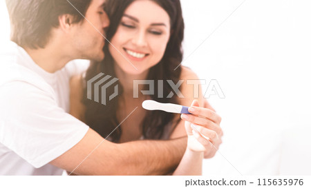 A young couple embraces with excitement as they share the good news of a positive pregnancy test. The woman holds the test up with a big smile, while the man leans in to kiss her on the cheek. 115635976