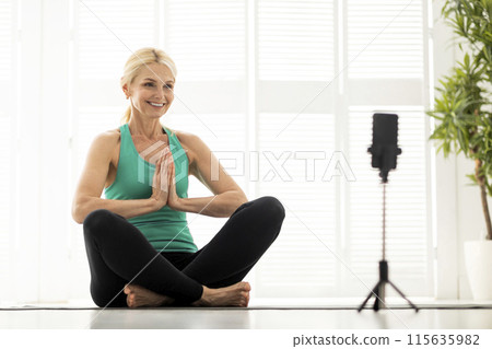 Online Yoga Class. Smiling Senior Woman Meditating In Front Of Smartphone At Home, Sporty Mature Female Sitting In Lotus Pose, Capturing Content For Social Media, Or Attending Virtual Training 115635982